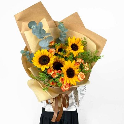 Sunflower bouquet with orange carnations and eucalyptus leaves, wrapped in brown paper.