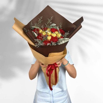 Chocolate and red carnation bouquet wrapped in brown paper, held by a person.