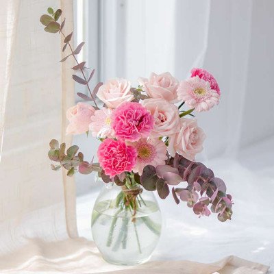 Pink roses, carnations, and gerbera daisies in a round glass vase with eucalyptus branches.