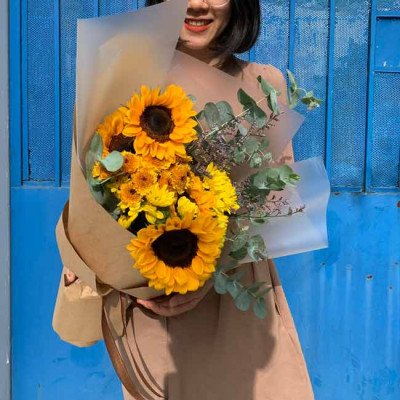 Woman holding a bouquet of sunflowers and yellow chrysanthemums wrapped in brown paper.