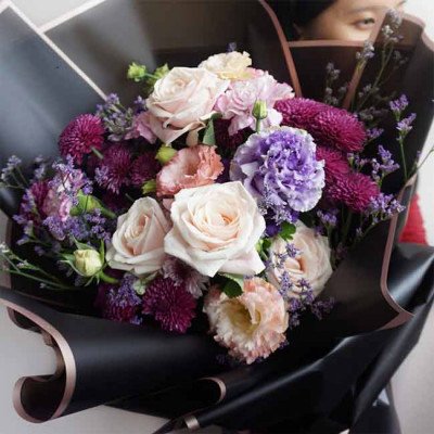 A bouquet of roses, carnations, and lisianthus in shades of pink, purple, and white, wrapped in black paper.