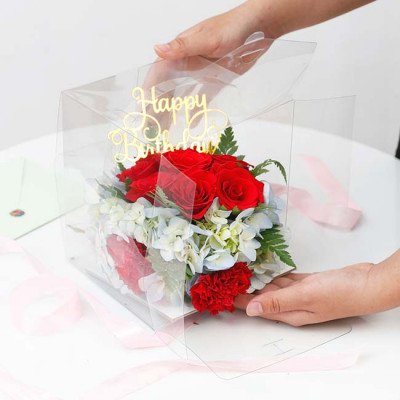 A clear box with red roses, red carnations, and blue hydrangeas, topped with a "Happy Birthday" banner.