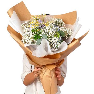 A person holds a bouquet of white baby's breath, yellow daisies, and blue flowers wrapped in brown paper.
