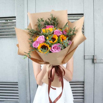 Bouquet of sunflowers and purple roses wrapped in brown paper, tied with a brown ribbon.