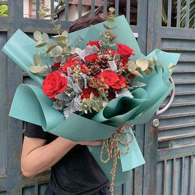 Bouquet of red roses, carnations, and pine cones wrapped in teal paper with a gold tie.