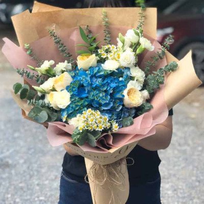 Bouquet of blue hydrangeas, white roses, and small white flowers wrapped in brown paper.