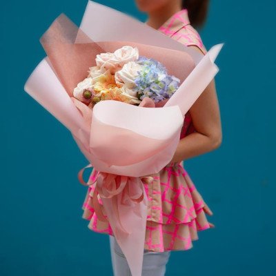 A woman holds a bouquet of pink roses, blue hydrangeas, and yellow flowers wrapped in light pink paper.