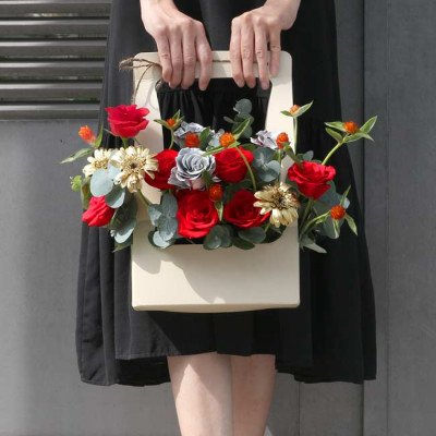A cream tote bag filled with red roses, grey roses, and daisies is held by a person in a black dress.