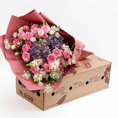 A bouquet of pink roses, purple hydrangeas, and other flowers in pink wrapping, displayed on a brown box.