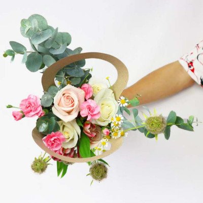 A hand holds a bouquet of pink and white roses, carnations, and daisies in a brown "8" shaped vase.