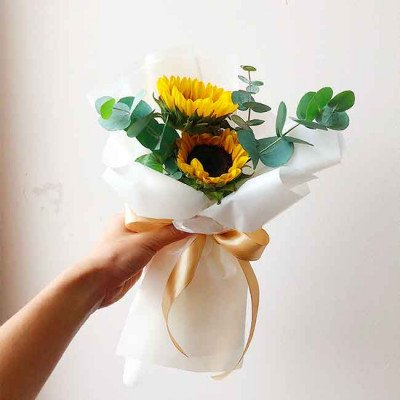 A hand holds a small bouquet of sunflowers and eucalyptus, wrapped in white paper and tied with a gold ribbon.