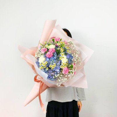 A person holds a large bouquet of pink, blue, and white flowers wrapped in pink paper.