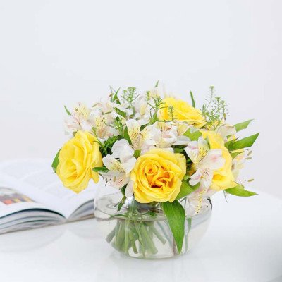 Yellow roses and white alstroemeria in a round glass vase on a white surface, with an open book to the left.