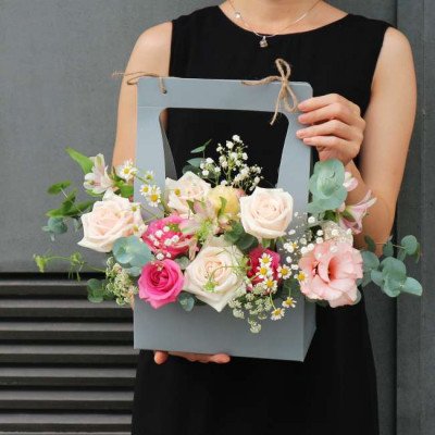 A person holds a pastel flower arrangement in a gray box, featuring roses, lisianthus, and baby's breath.