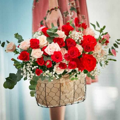 A hand holding a basket of red and pink roses with greenery.