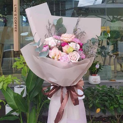 Bouquet of pink and purple carnations, roses, and eustoma flowers with greenery, wrapped in beige paper and tied with a brown ribbon.