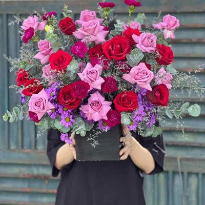 Pastel purple and red rose floral arrangement in a black box held by a person in a black dress.