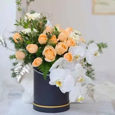 Peach roses and white orchids in a black box, with silver-dusted ferns.