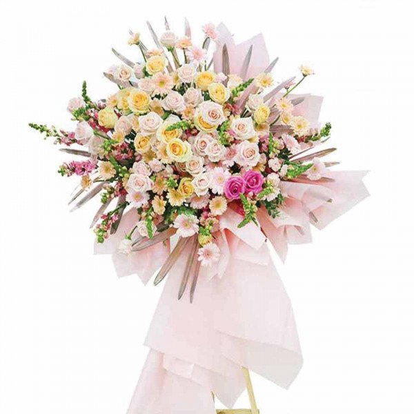 A grand floral arrangement of pink and yellow roses, gerbera daisies, and greenery, displayed on a stand with a pink wrap.