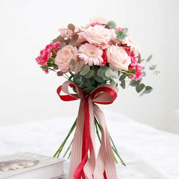 Bridal bouquet of pink roses, gerbera daisies, and carnations with eucalyptus, tied with a red and pink ribbon.