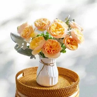 A bouquet of peach and orange roses in a white vase with a jute tie, on a round woven tray.