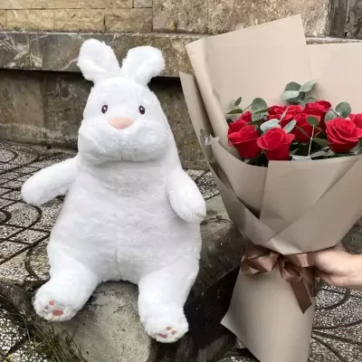 Fluffy white rabbit plush toy with pink accents sits next to a bouquet of red roses.
