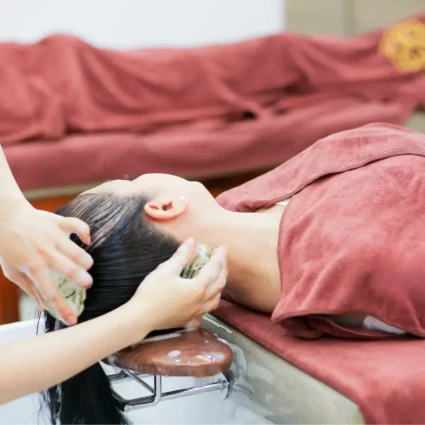 Person receiving a herbal hair wash with green herbal massage balls on a massage table.