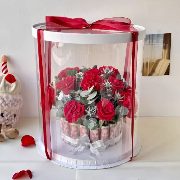 Money flower arrangement in a clear box with red ribbon, featuring red roses and eucalyptus.