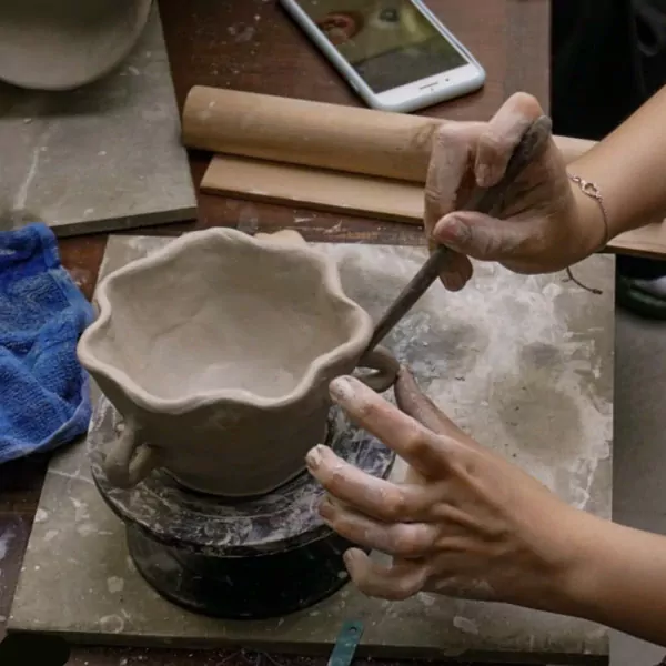 Hands shaping a clay bowl with a tool in a pottery workshop.