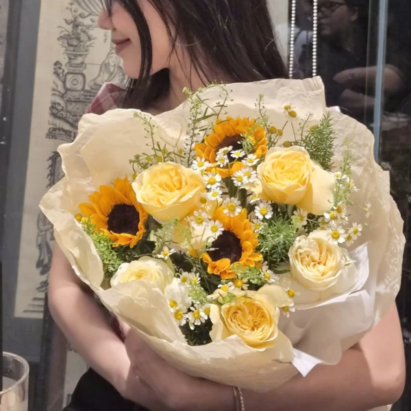 A bouquet of sunflowers, yellow roses, and small white flowers, wrapped in light brown paper.