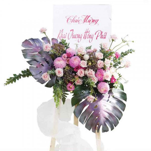 Pink and white roses, chrysanthemums, and tropical leaves on a floral stand with a banner.
