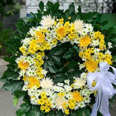 Wreath of yellow and white flowers with greenery and a white ribbon.