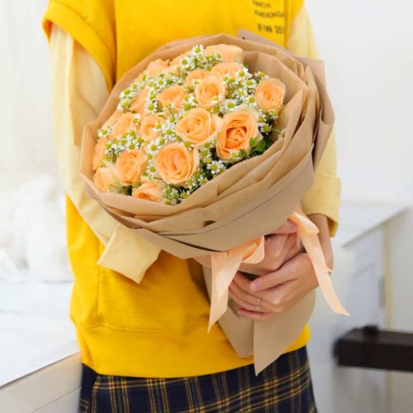 Peach roses and white daisies in a brown paper bouquet held by a person in a yellow sweatshirt.