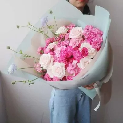 A lush bouquet of pink roses and carnations with delicate purple flowers, wrapped in pale blue paper.