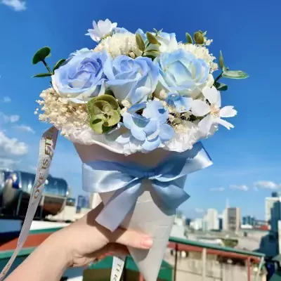 A cone-shaped bouquet of pastel blue and white roses, tied with a blue ribbon, held against a bright blue sky.