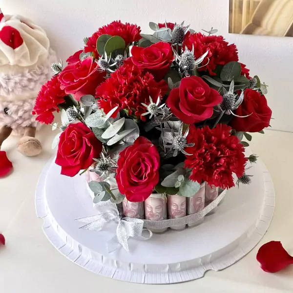 Red roses, carnations, and thorny thistles in a money cake box, with a stuffed toy nearby.