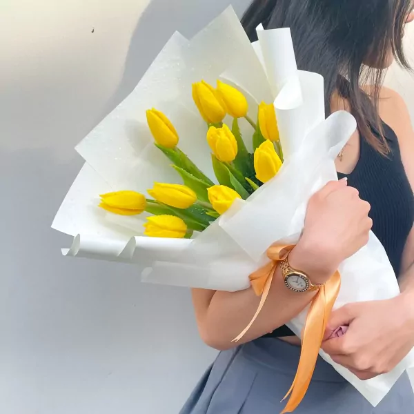 Bouquet of yellow tulips with white wrapping and an orange ribbon, held by a woman.