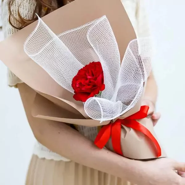 A hand holds a red rose bouquet wrapped in brown paper and white mesh, tied with a red ribbon.