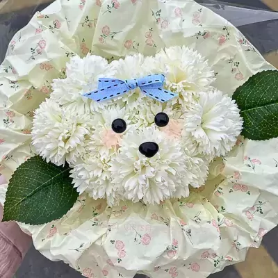 White flower arrangement shaped like a puppy's face with a blue bow, on floral wrapping paper.