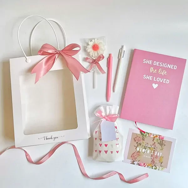 Pink and white gift set for women including a notebook, pens, a flower lollipop, a candle, and a "Happy Women's Day" card.