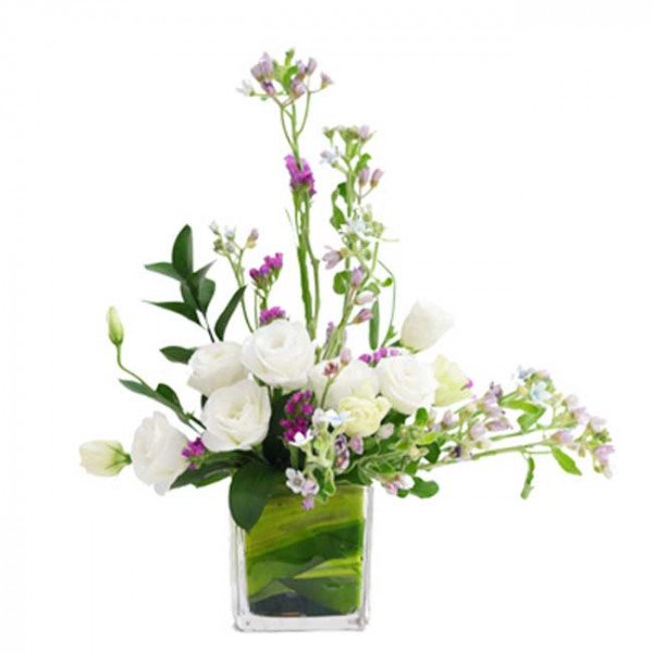 White roses, statice, and blue star flowers arranged in a square glass vase with green foliage.