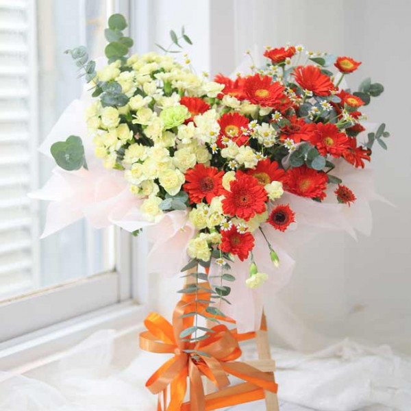 Bouquet of red gerberas, white carnations, and eucalyptus leaves on a wooden stand with an orange ribbon.