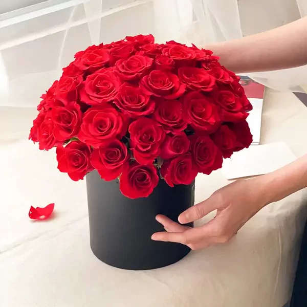 A black box overflowing with vibrant red roses, a single rose petal lies to the side.