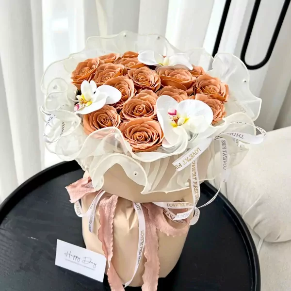 Peach roses and white orchids in a bouquet, wrapped in sheer fabric and paper, with a "Happy Day" card.