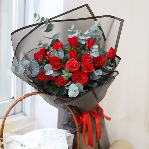 Bouquet of red roses and eucalyptus, wrapped in black paper with a red ribbon, in a basket with a card.
