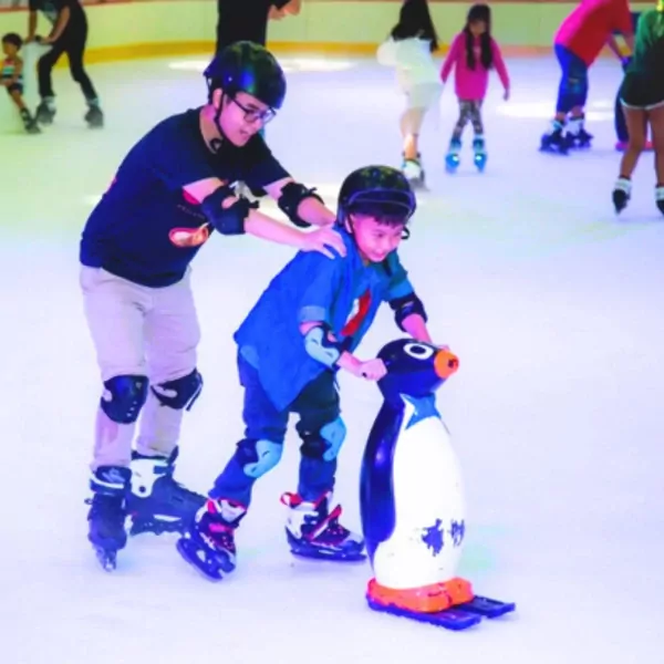 Man in helmet helps child in helmet on ice skates with penguin.