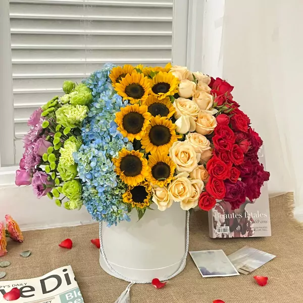 Round flower box with rainbow arrangement of sunflowers, hydrangeas, and roses in red, cream, and purple.