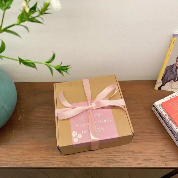 A gift box with a pink ribbon, on a wooden table with books and a vase of flowers.