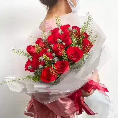 A bouquet of red roses with greenery and a red ribbon, held by a person in a pink dress.