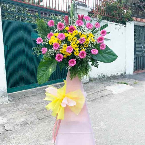 A tall floral arrangement of pink and yellow flowers with a pink conical base and yellow bow.